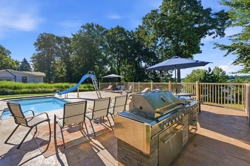 Backyard patio with outdoor kitchen, dining set and in-ground pool surrounded by trees